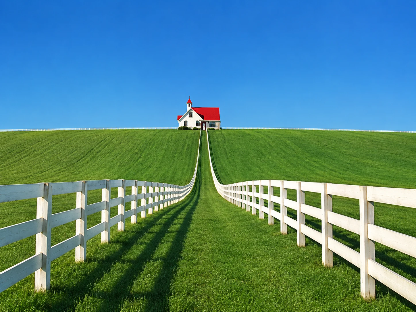 A white house with a red roof beyond green fields, with white fences converging toward it.